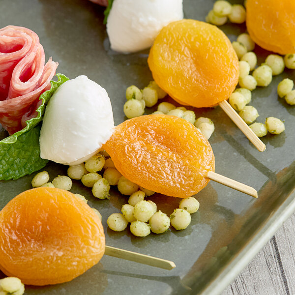A plate with skewered dried apricots, mozzarella balls, and small pieces of salami, garnished with herbs and small round snacks.