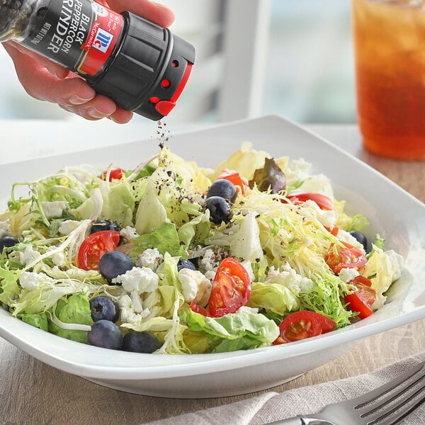 A hand using a McCormick Black Peppercorn Grinder on a salad on a table.