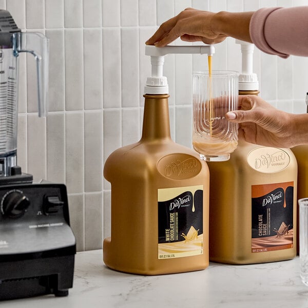 A large gold bottle of DaVinci Gourmet White Chocolate Flavoring Sauce with a pump dispenser being used to pour sauce into a glass.