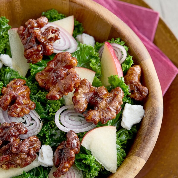 A wooden bowl filled with a salad containing raw walnut halves and pieces, kale, red onion slices, apple slices, and crumbled cheese.