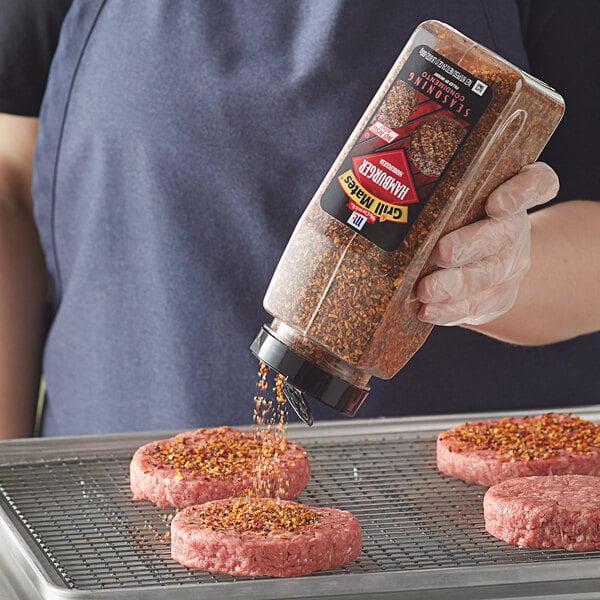 A person pouring McCormick Grill Mates Hamburger Seasoning onto a hamburger patty.