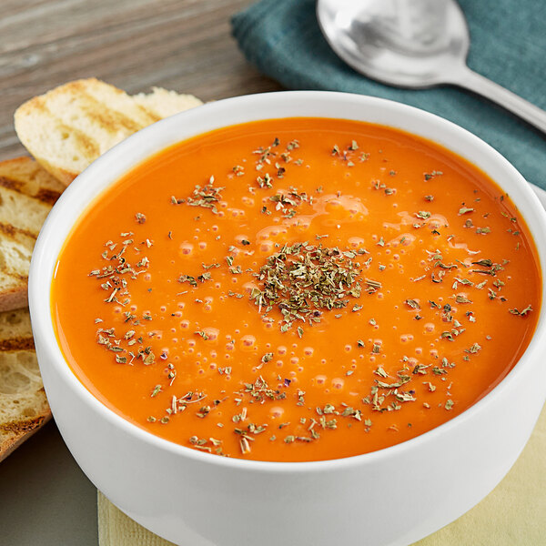 A bowl of orange soup with McCormick basil leaves on top next to a spoon and toast.