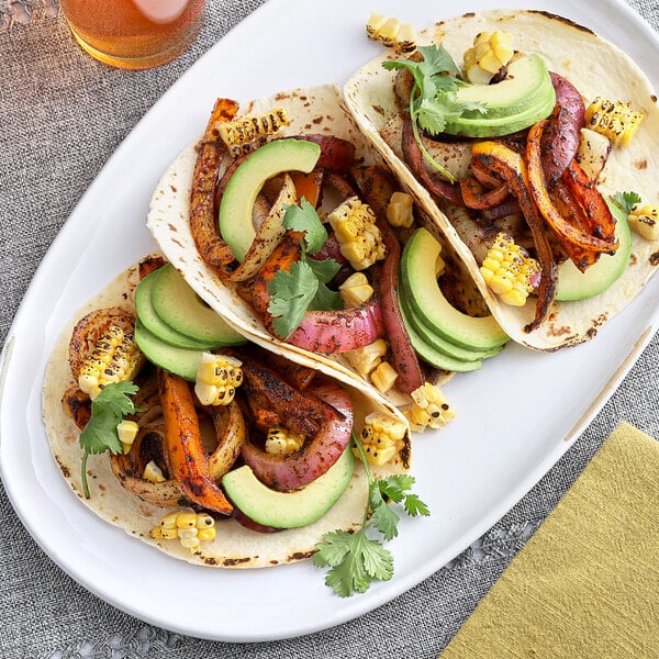 A white plate with a taco filled with avocado and corn on a tortilla with a plate of grilled vegetable and corn tacos.
