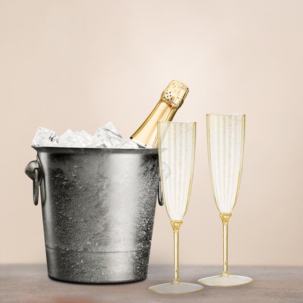 A Posh Setting gold plastic champagne flute filled with champagne next to a bottle in a bucket of ice.