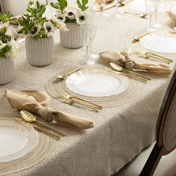 A table set with white Posh Setting clear plastic plates with gold bands and gold silverware, with white flowers in a vase.