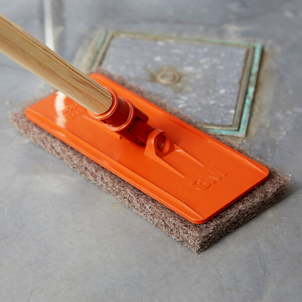 A 3M Doodlebug brown scrub pad with a wooden handle on a surface.