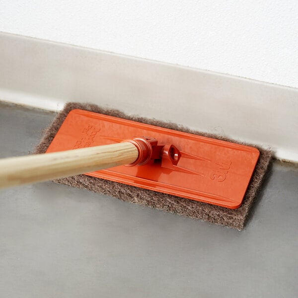 A person using a 3M Doodlebug brown heavy-duty scrub pad to clean a floor.