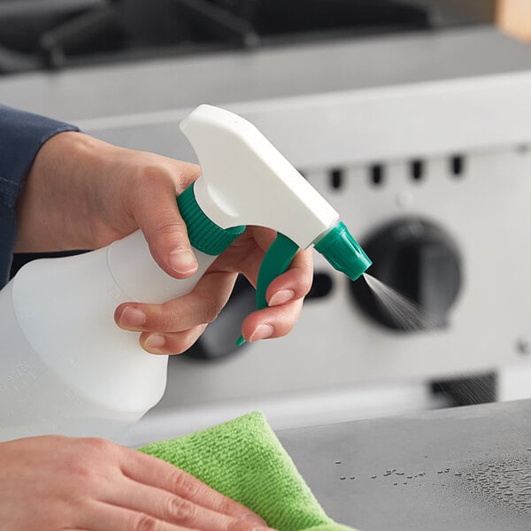 A person using a Lavex green plastic spray bottle to clean a counter in a professional kitchen.