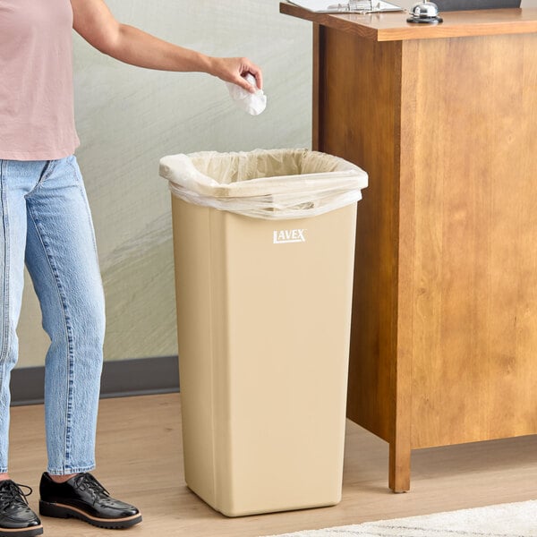 A beige square trash can with a plastic liner, labeled 'Lavex,' placed next to a wooden counter.