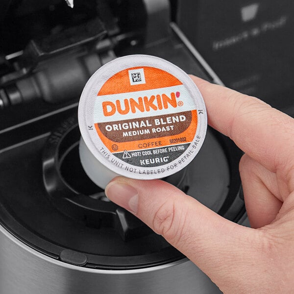 A hand holding a small round container of Dunkin' Original Blend coffee pods.