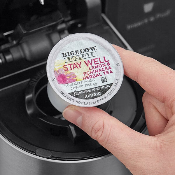 A hand holding a small white container of Bigelow Benefits Lemon and Echinacea Herbal Tea K-Cup Pods with a label.