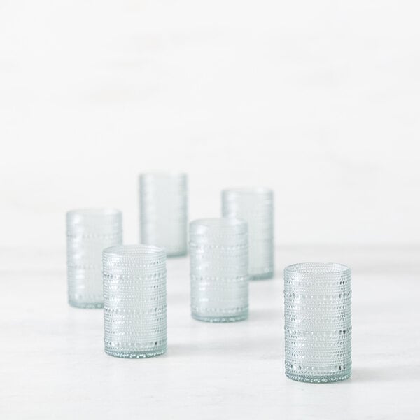 Six textured, cylindrical, light green drinking glasses are arranged on a white surface with a soft, minimal background.