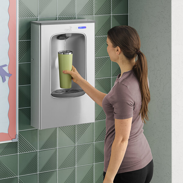 A stainless steel contactless bottle filling station mounted on a tiled wall, with a person filling a reusable bottle.