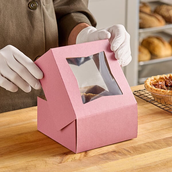 A person in gloves opening a pink Baker's Lane box with a clear window to reveal a pastry.