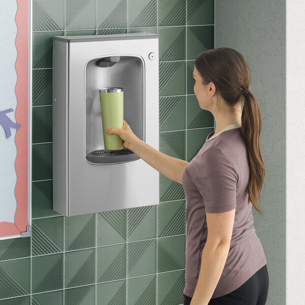 A woman filling a reusable bottle at a stainless steel wall-mounted bottle filling station.