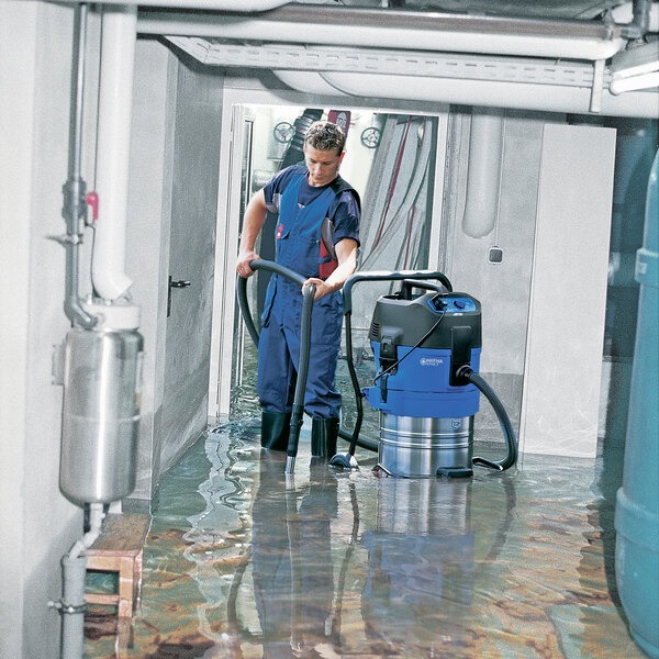 A man in blue work clothes using a Nilfisk wet/dry vacuum to clean a flooded floor.