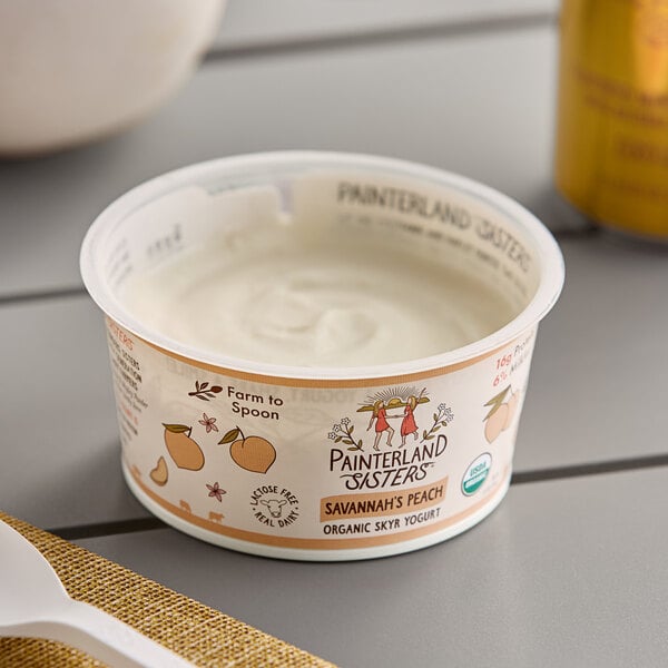 A white Painterland Sisters Savannah's Peach Organic Skyr Yogurt container with a spoon on a table.