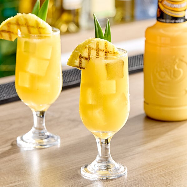 Two glasses of yellow liquid with pineapple slices and green leaves on the side.
