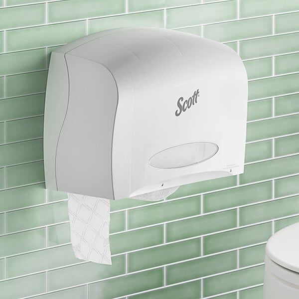 A white Scott Coreless Jumbo Roll toilet paper dispenser with a stub roll section on a green tile wall.