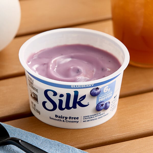 A plastic container of Silk blueberry soymilk yogurt alternative on a table.