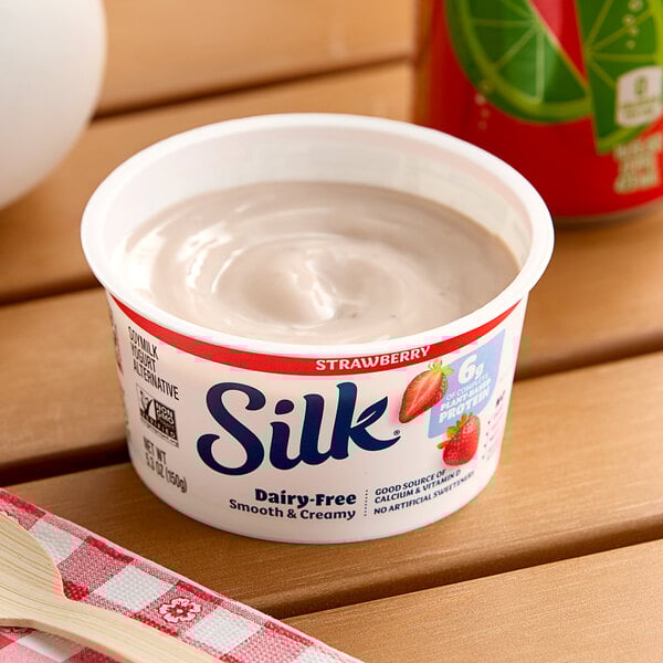 A container of Silk strawberry soymilk yogurt alternative on a table with a wooden spoon.