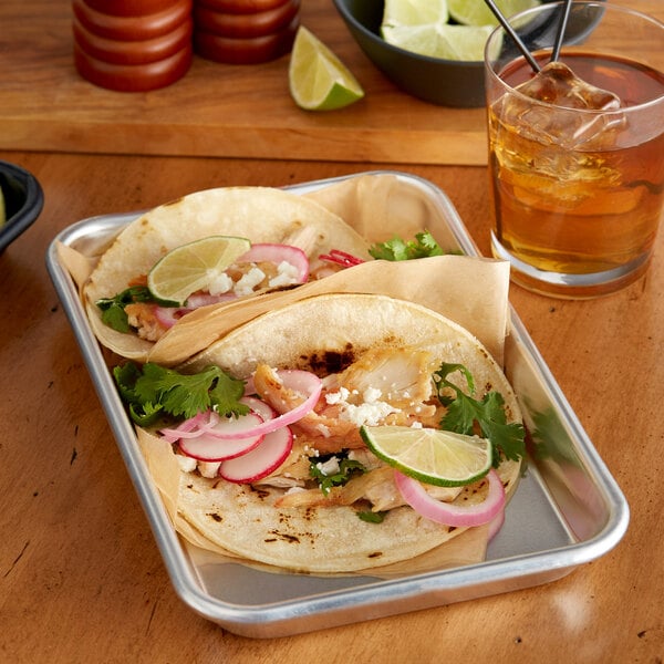 A tray of tacos with vegetables and meat on a table with a glass of amber liquid with ice and a straw.