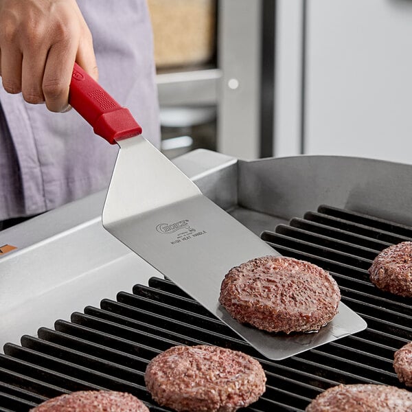 A hand using the Choice turner to cook a burger on a grill.