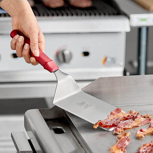 A hand using a Choice square edge turner with a red handle to lift bacon from a grill.