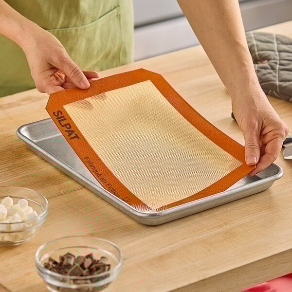 A person using a Sasa Demarle SILPAT® silicone baking mat on a cookie sheet filled with chocolate chunks.
