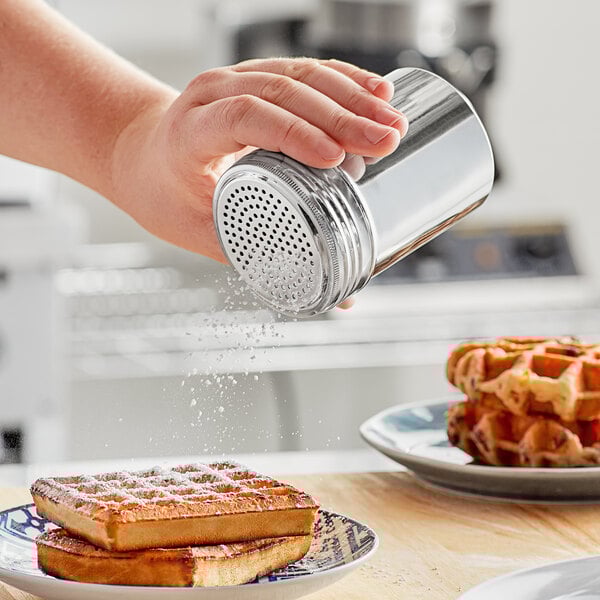 A person sprinkling powder on waffles from a silver Choice stainless steel shaker.