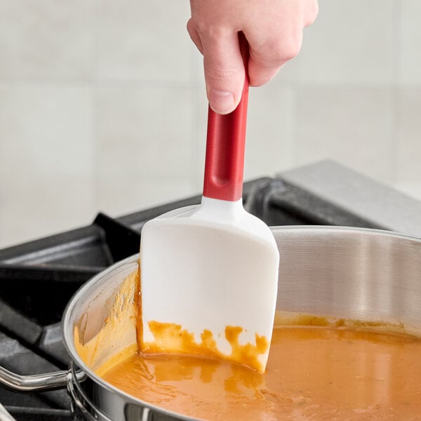A hand is using a white and red silicone spatula to stir orange sauce in a stainless steel pot on a stove.