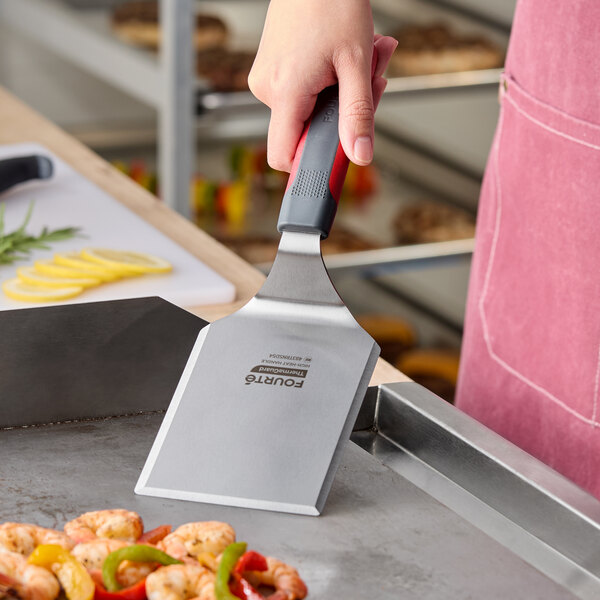 A stainless steel high heat solid beveled square edge turner with a nylon handle being used on a griddle.