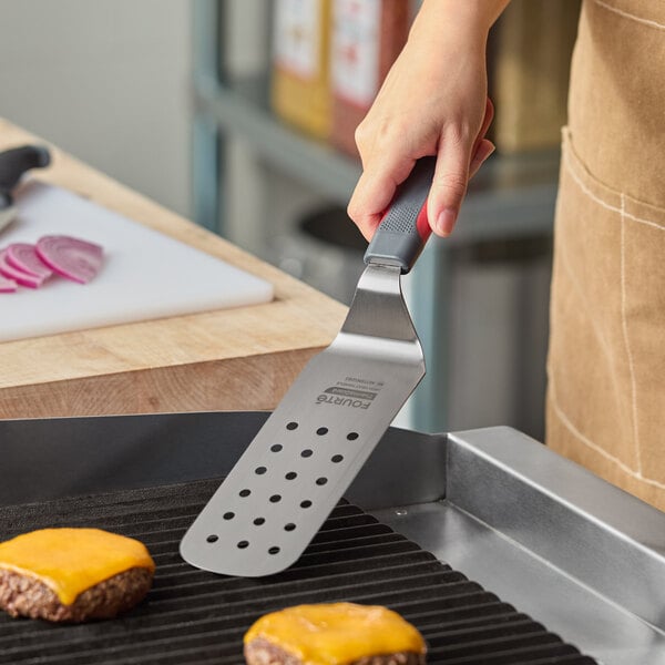 A stainless steel perforated turner with a nylon handle being used to cook cheeseburgers on a grill.