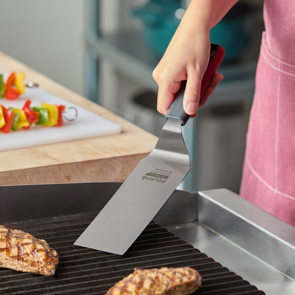 A stainless steel high heat solid square edge turner with a nylon handle being used to cook food on a grill.