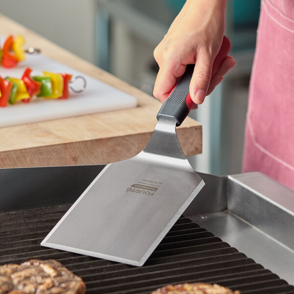 A stainless steel square edge turner with a black nylon handle being used on a grill.
