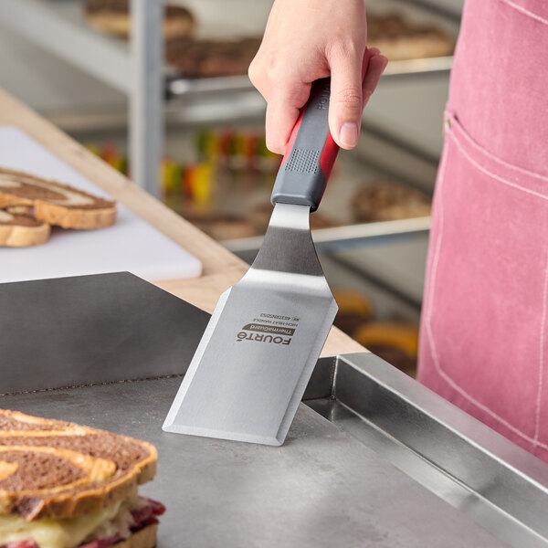 A stainless steel high heat solid beveled square edge turner with a nylon handle being used on a griddle.