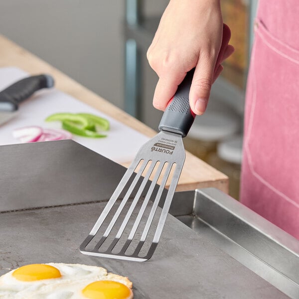 A stainless steel slotted fish and egg turner with a black nylon handle being used on a griddle.