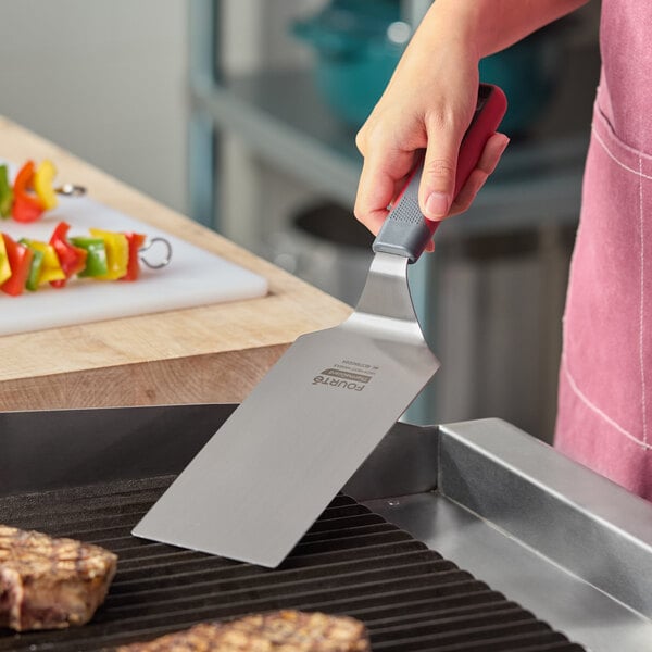 A stainless steel high heat solid square edge turner with a nylon handle being used on a grill.