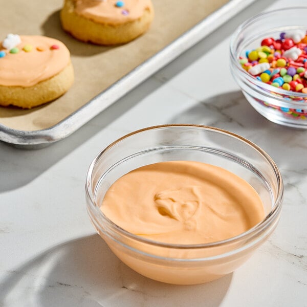 A glass bowl filled with orange royal icing mix on a countertop near decorated cookies and sprinkles.