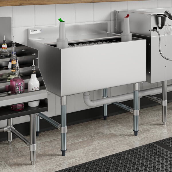 A Regency underbar ice bin machine on a counter with sliding lid open over ice and bottles on a shelf.