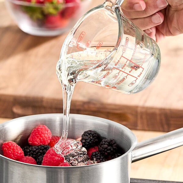 A clear liquid glucose syrup being poured from a measuring cup into a saucepan with berries.
