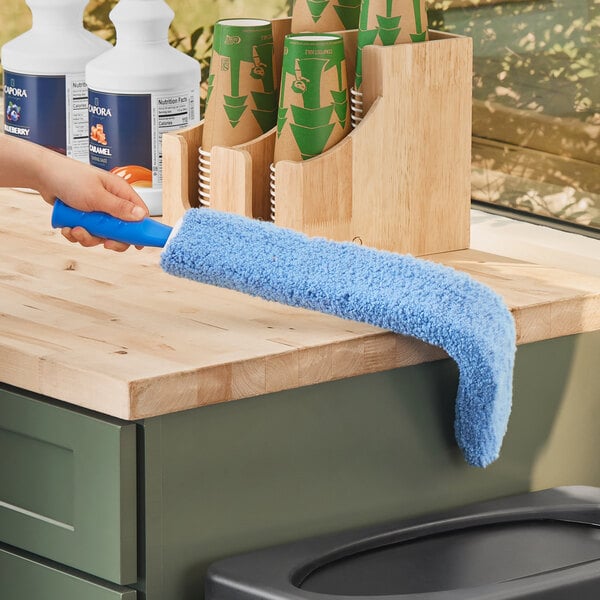 A hand using a Lavex blue duster with a black wand to clean a white surface.
