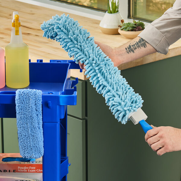 A blue Lavex 23-inch flex wand duster with a chenille sleeve being held near a cleaning cart.