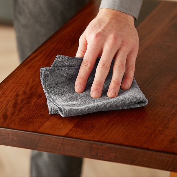 A hand using a Lavex charcoal gray microfiber cloth to wipe a table.