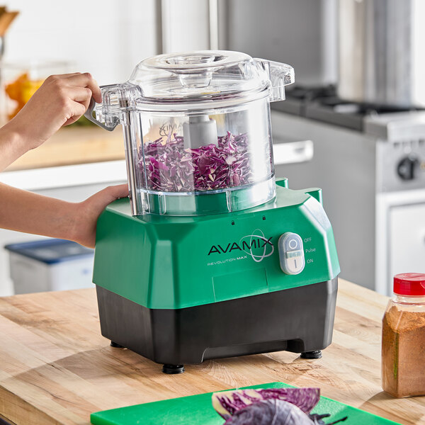 A green and black AvaMix Revolution Max food processor with a clear plastic batch bowl, shown processing chopped purple cabbage on a kitchen counter.