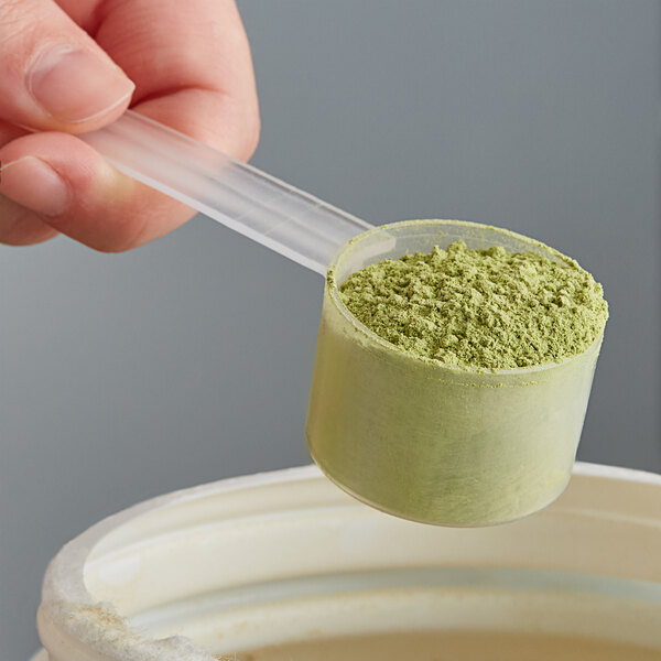 A hand holding a long-handled plastic polypropylene scoop over a container of green powder.