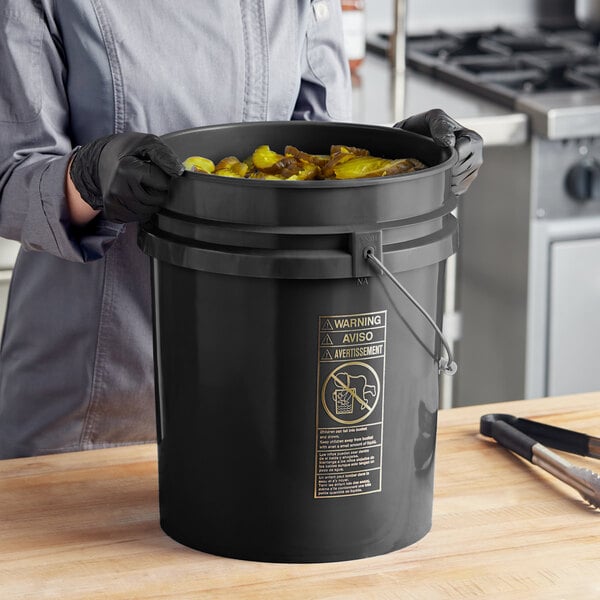 A woman in gloves holding a BWAY black round pail with a metal handle full of food.
