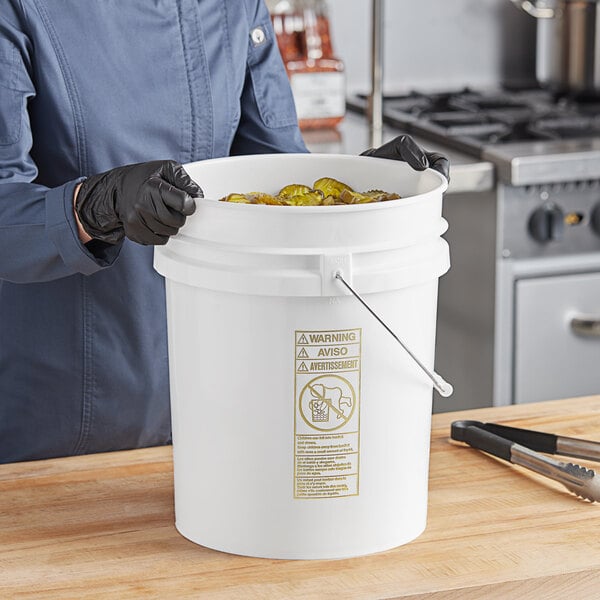 A person wearing black gloves holding a white BWAY pail full of food.
