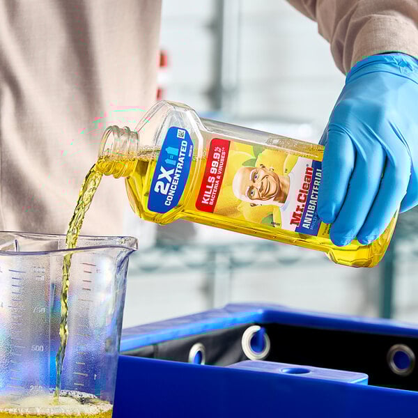 A hand in a blue glove pouring Mr. Clean Antibacterial Multi-Surface Cleaner into a measuring cup.