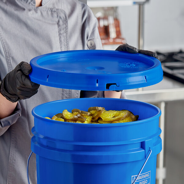 A woman in a chef's uniform holding a blue BWAY round pail cover over a blue bucket full of pickles.
